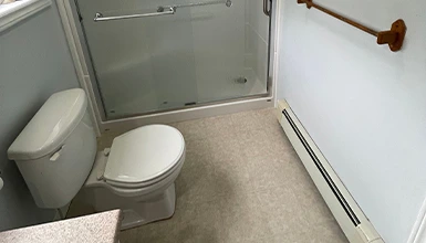 Bathroom with toilet, glass shower door, baseboard heater, and towel rack on a light-colored floor.