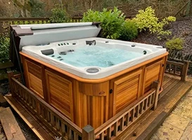 Outdoor hot tub with wood siding and open cover, surrounded by greenery.